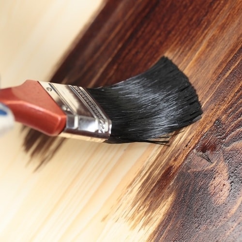 Brown wooden handle paint brush with black bristles painting a dark brown wood stain on a light tan piece of wood