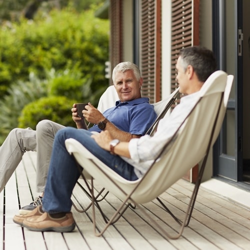 Two men in cloth chairs sitting and talking while out on a eggshell-colored back deck flooring with brown door and shutters
