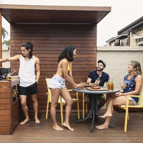 Three people talking around table outdoors while one person cooks food, all surrounded around wooden shelter