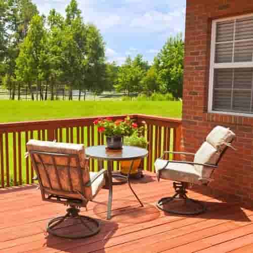 Outdoor sitting area with padded chairs around a small table with a potted plant on a brown wooden porch connected to a red bricked wall and window surrounded by grass and trees in the distance 