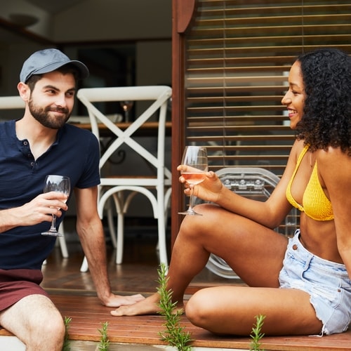 Man and woman holding wine glasses smiling and looking at each other sitting on brown wooden flooring in front of white chairs