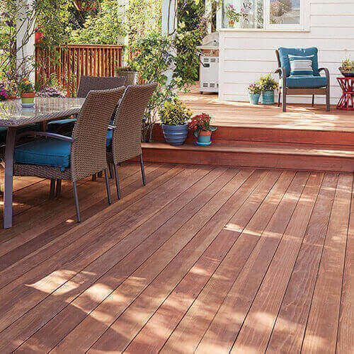 Outdoor brown wooden deck with blue padded wooden chairs and potted plants with white house in background and trees