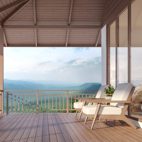 Outdoor fenced porch with light brown wooden panel floors and ceiling with large windows to inside, padded wooden chairs, and mountains in background