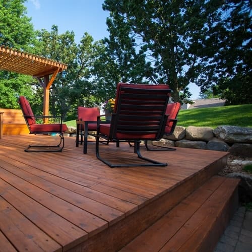 Outdoor patio with wooden floor with red padded metal chairs and wooden fence roofing above surrounded by trees with blue sky