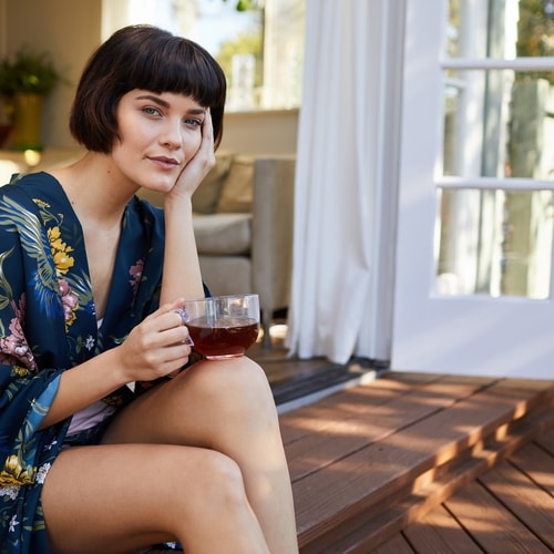 Woman with dark short hair leaning her head on her hand looking into camera holding a glass of tea sitting on steps with white door and living room in background 