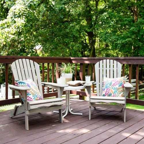 Two white wooden chairs next to table with potted plant and accessories on a wooden fenced in deck surrounded by trees