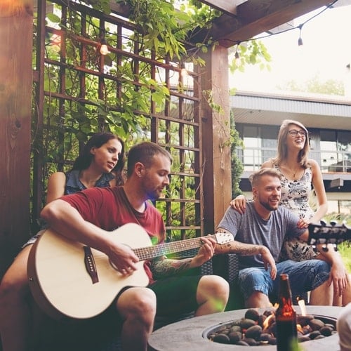 Two women and two men sitting while one is playing guitar in roofed wooden gazebo covered in vines with house in background