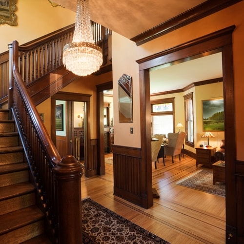 Inside house with light tan walls with brown accents and frames with hanging chandelier, mirror, paintings, windows, and distant furniture